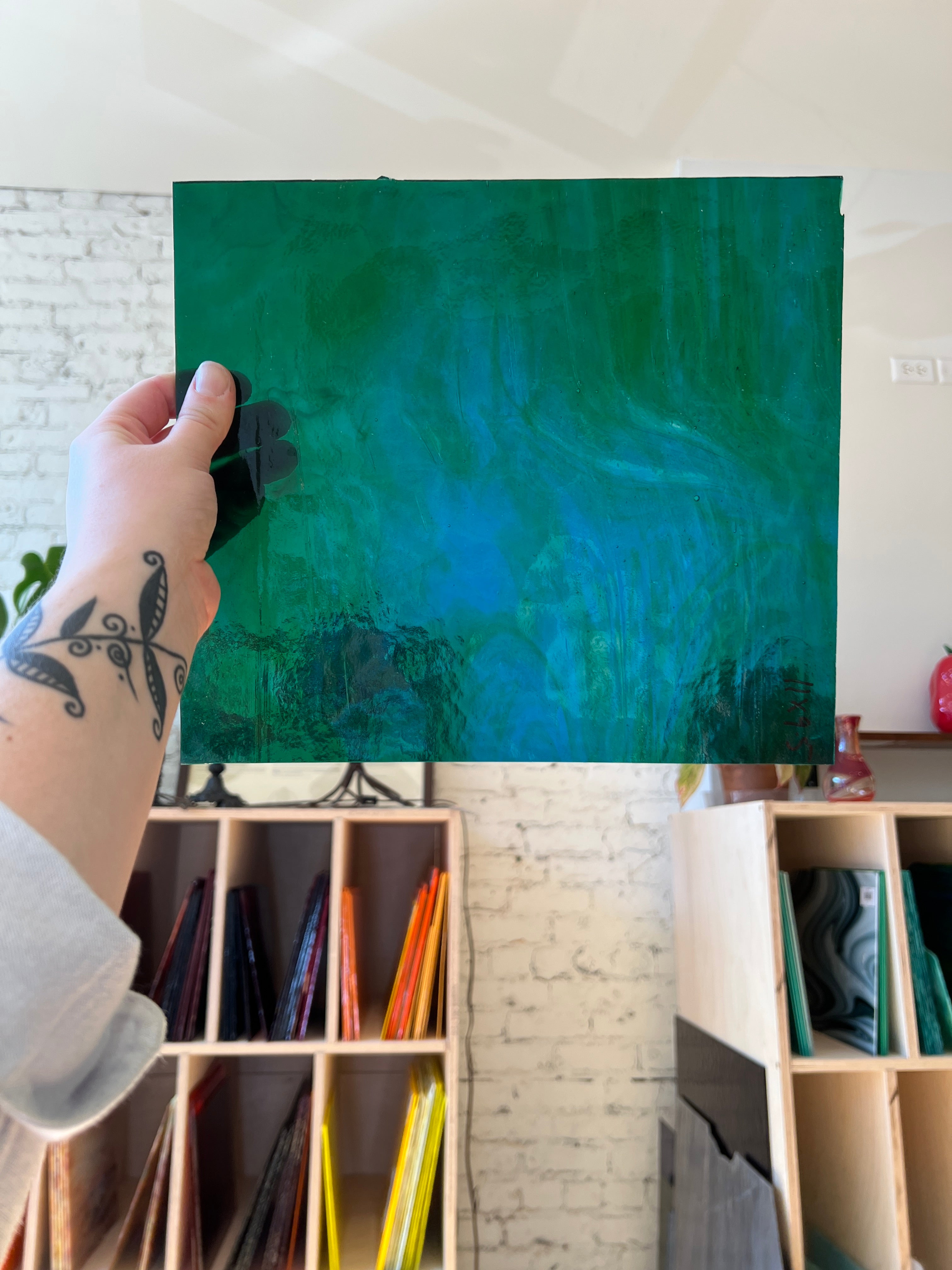 Person holding a green and blue swirled textured piece of stained glass in front of a blurred sunny interior. 