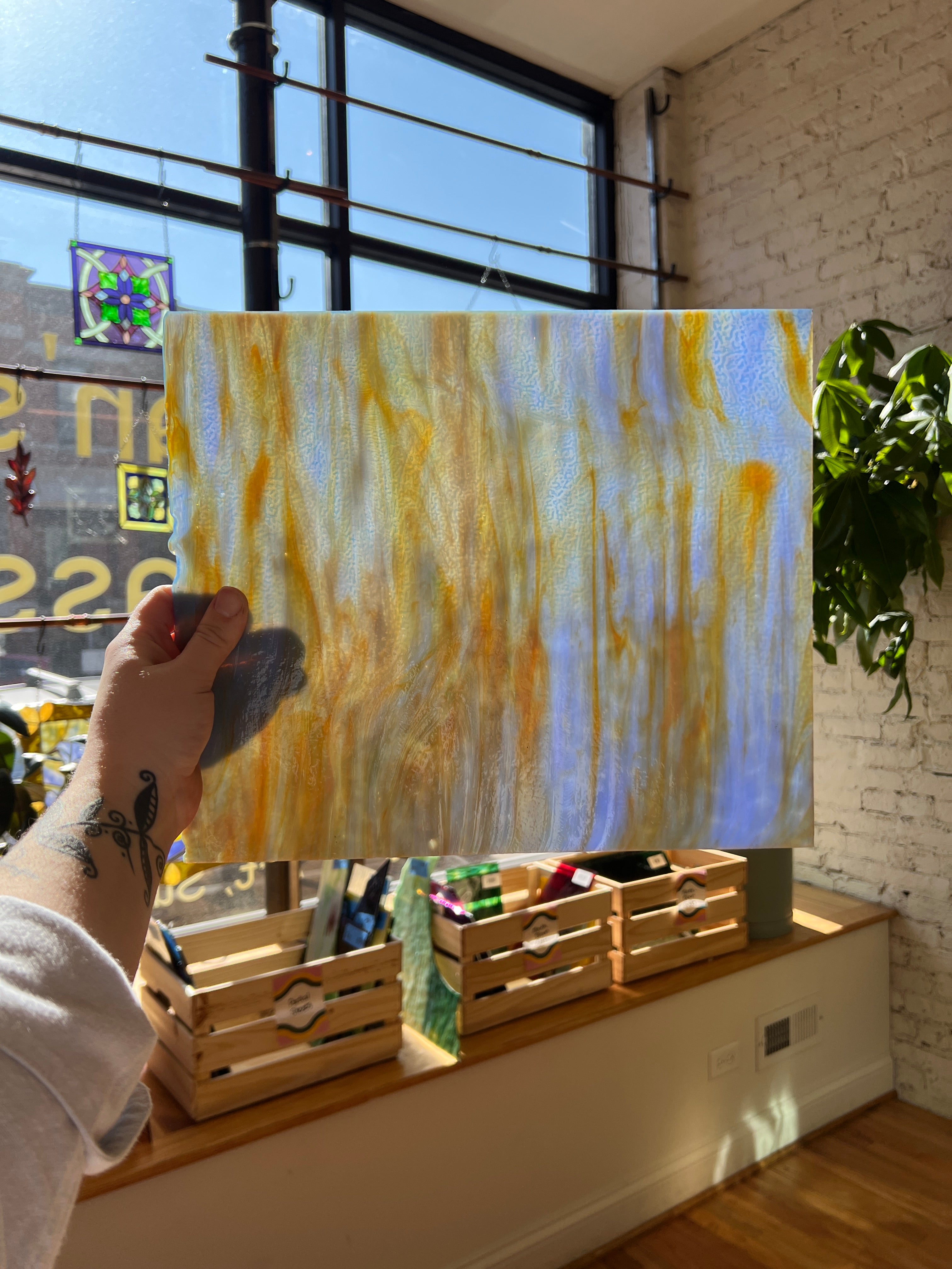 Person holding a transparent and gold swirled piece of stained glass in front of a blurred sunny window. 