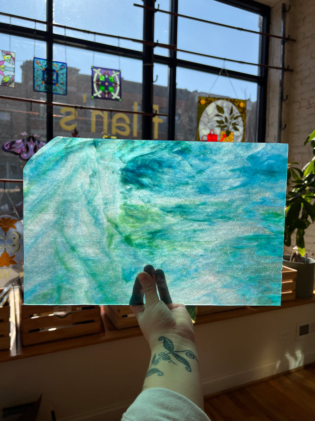Person holding a blue and green swirled piece of stained glass in front of a blurred sunny window.