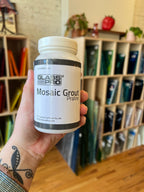 Hand holding a bottle of Glass Pro Mosaic Grout Praline in front of an indoor background.