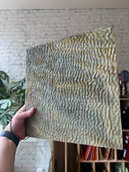 Hand holding a textured brown panel of stained glass with anwavy pattern against a blurred indoor background
