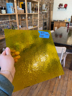 Person holding a gold-colored sheet of glass with a blue label in a room with shelves and furniture.