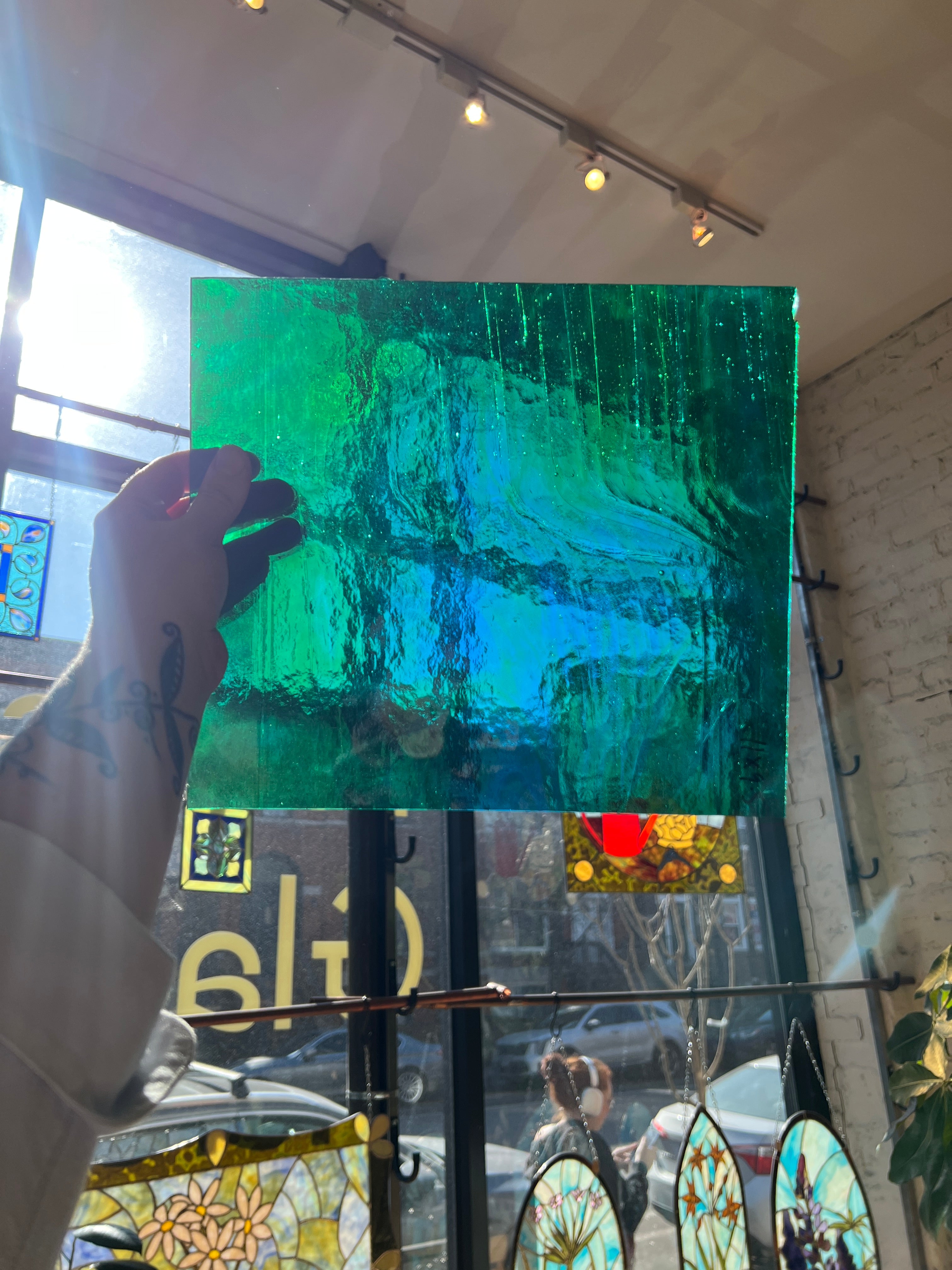Person holding a green and blue textured piece of stained glass in front of a blurred sunny window. 