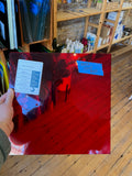 Red glass piece of glass with a label held by a hand, on a wooden floor with shelves in the background.