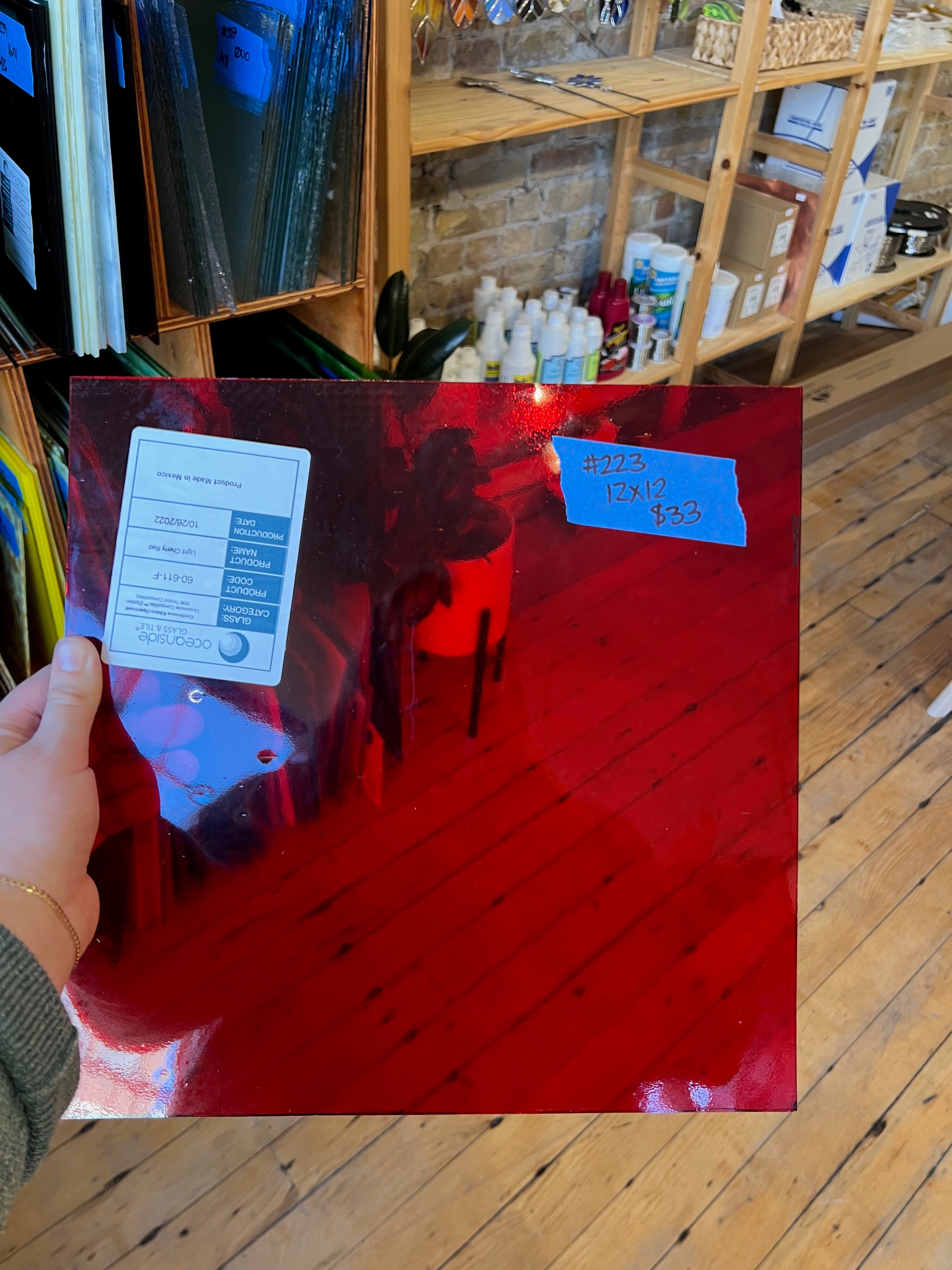 Red glass piece of glass with a label held by a hand, on a wooden floor with shelves in the background.