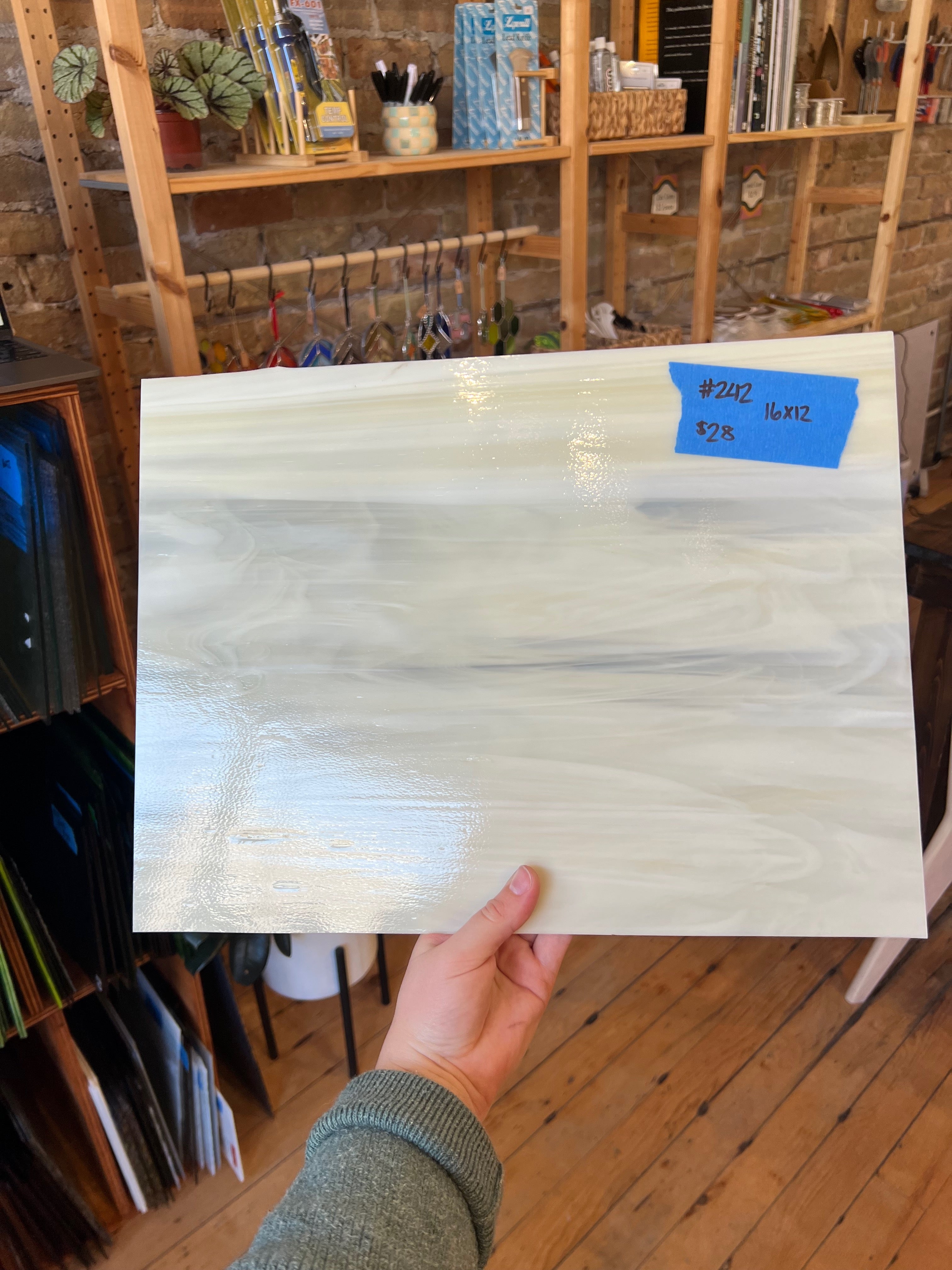 Person holding a large white sheet of glass with blue tape in a room with shelves and plants.