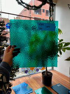 Person holding a piece of teal textured glass in a room with plants and a window.