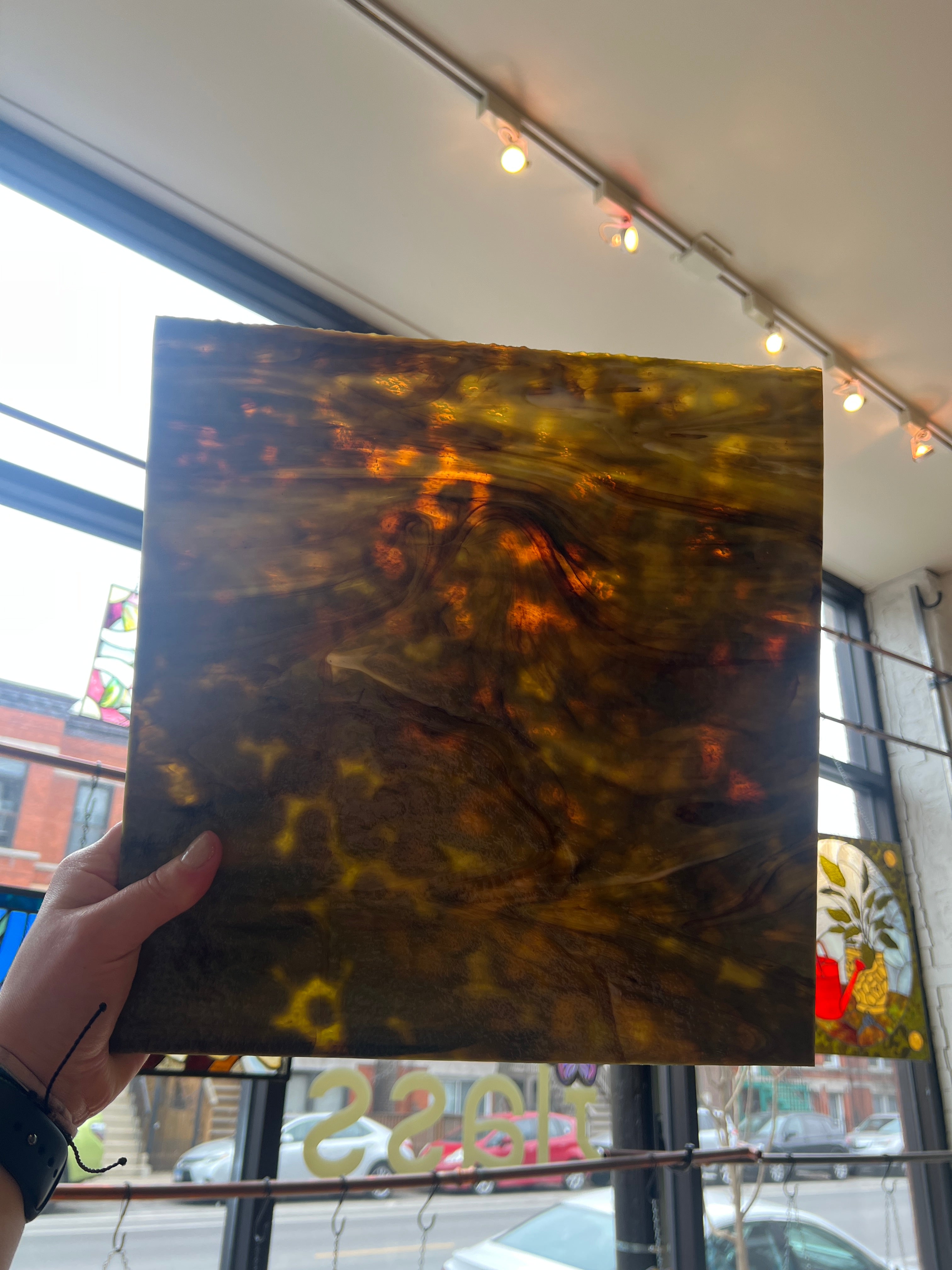 Hand holding a square piece of stained glass with abstract design in front of a window