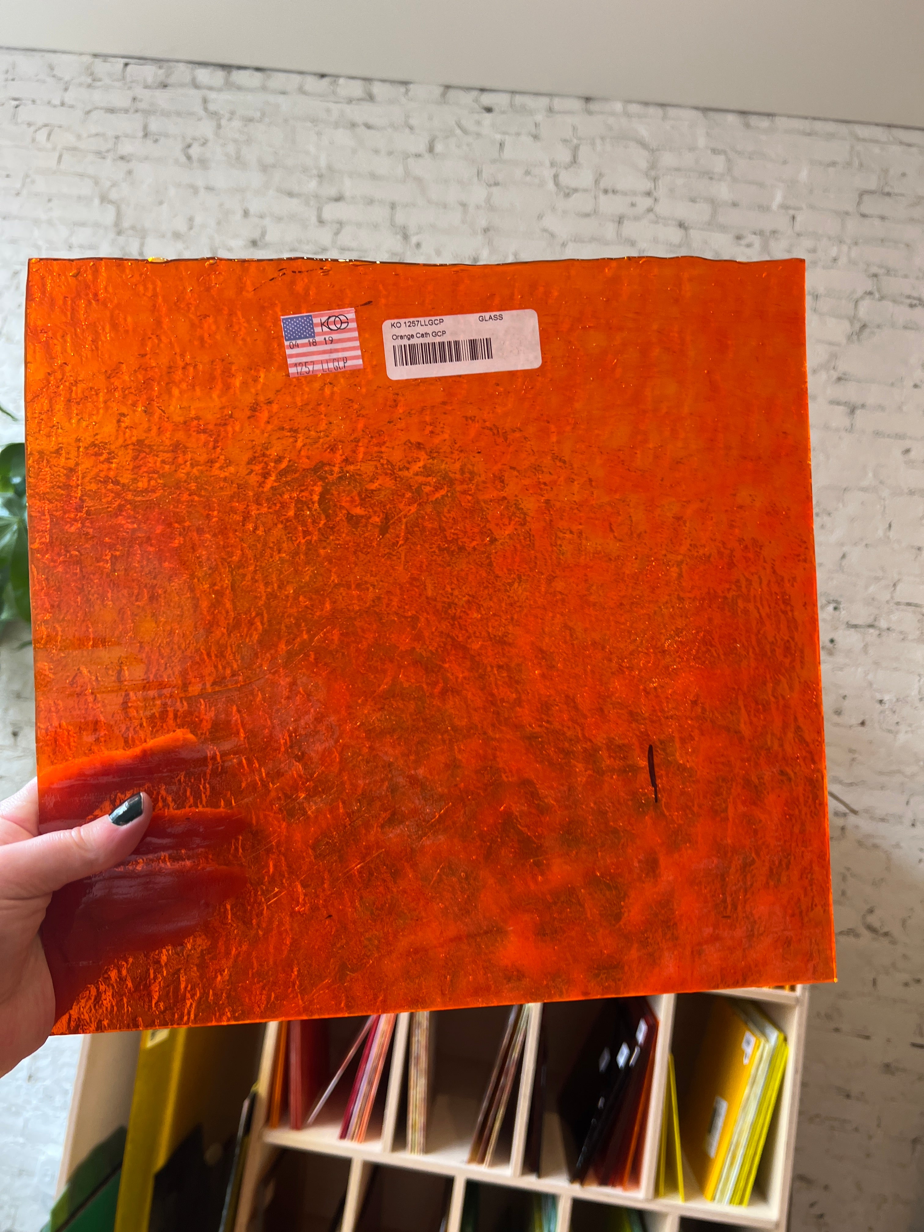 Orange piece of stained glass with a barcode and label held by a hand, on a textured surface.