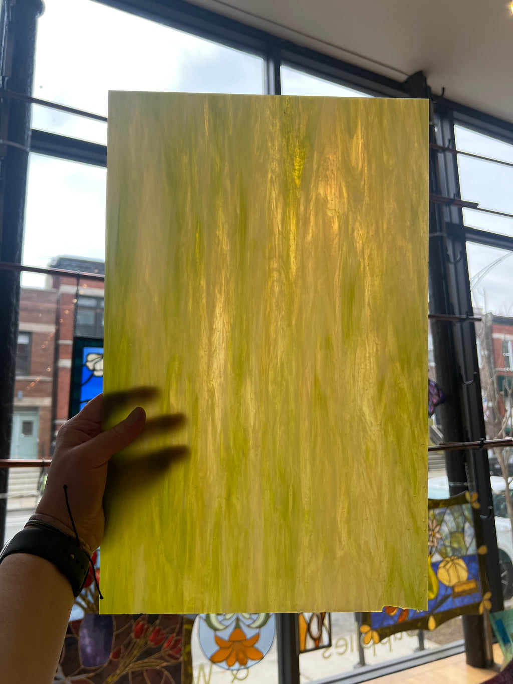 Hand holding a large rectangular yellow sheet of stained glass with a blurred indoor background