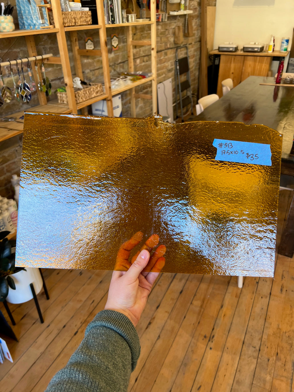 Hand holding a gold-colored sheet of glass in an indoor setting with wooden shelves and furniture.