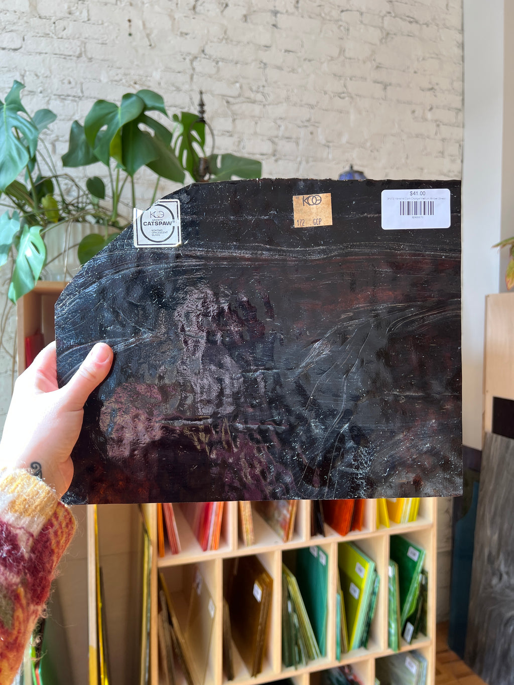 Person holding a black textured piece of stained glass with a brand label, indoor setting with plants and shelves.