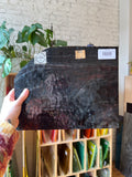 Person holding a black textured piece of stained glass with a brand label, indoor setting with plants and shelves.