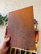Hand holding a textured brown sheet of stained glass in front of a colorful shelf.