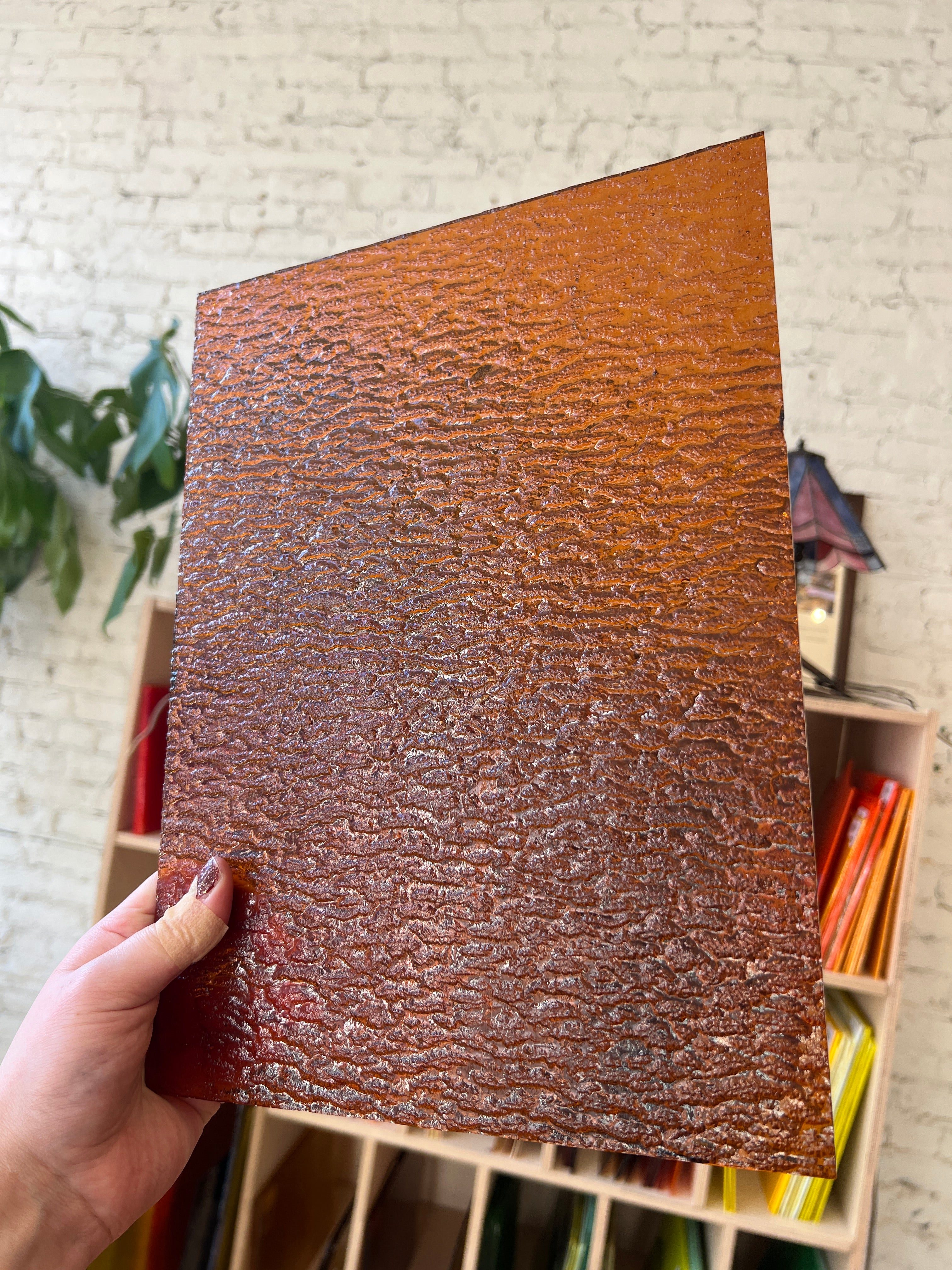Hand holding a textured brown sheet of stained glass in front of a colorful shelf.