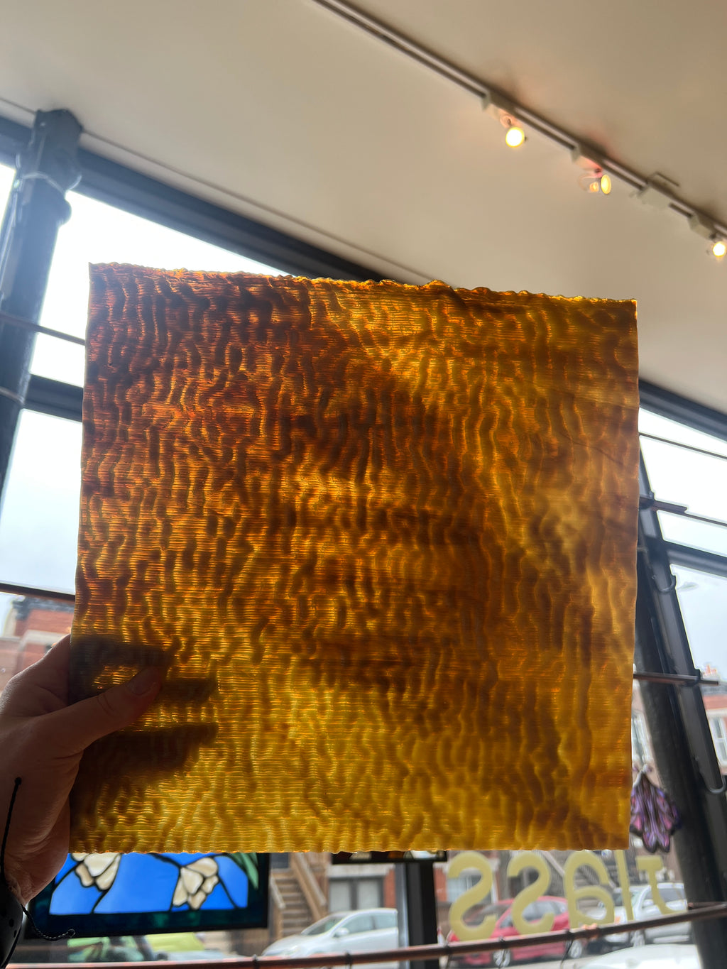 Hand holding a square piece of brown stained glass with a wavy texture in front of a window.