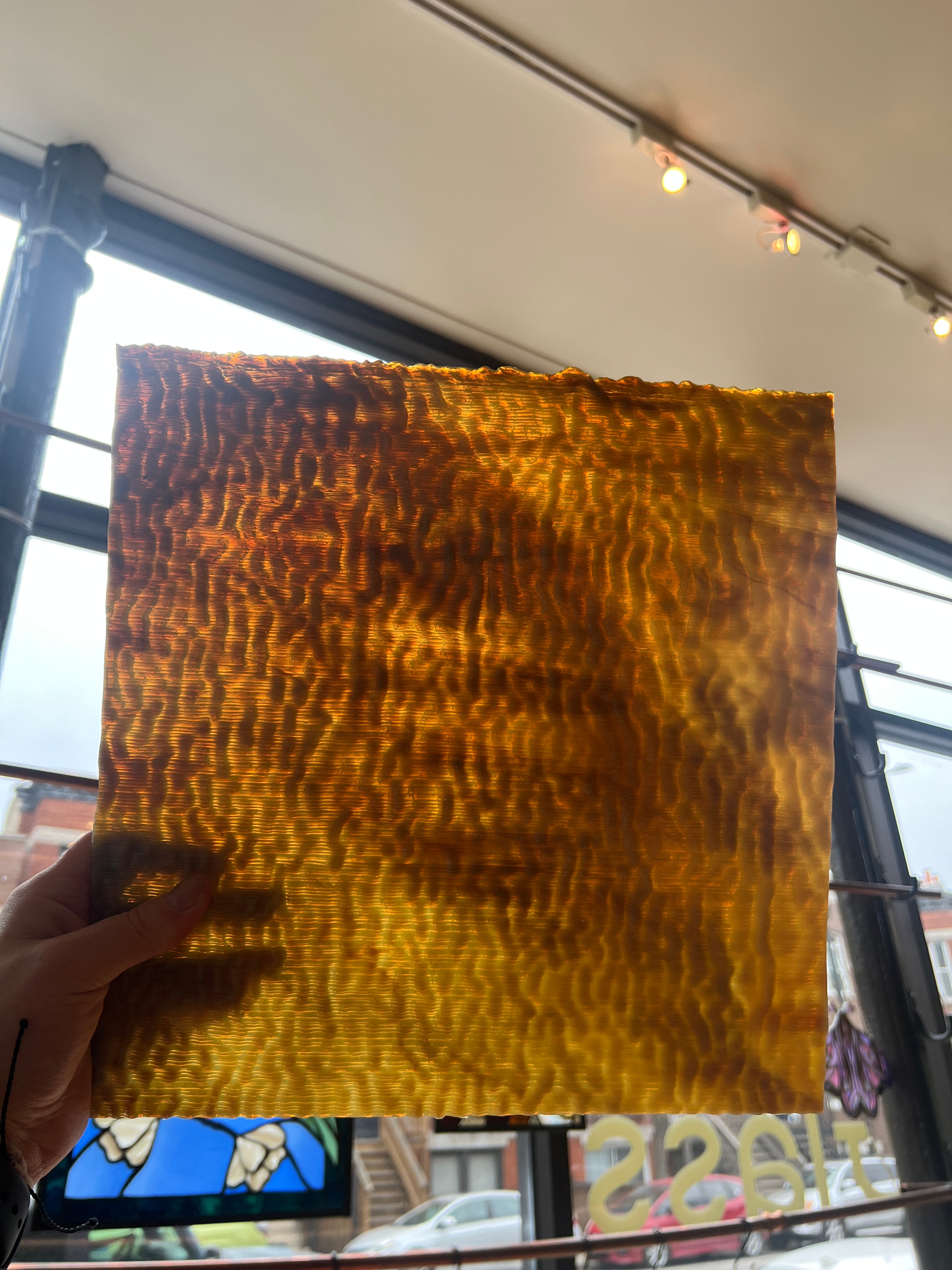 Hand holding a square piece of brown stained glass with a wavy texture in front of a window.