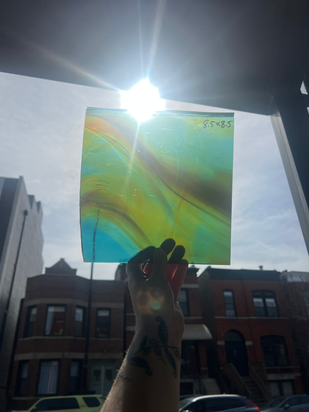 Hand holding a colorful abstract piece of stained glass in front of a window with buildings outside