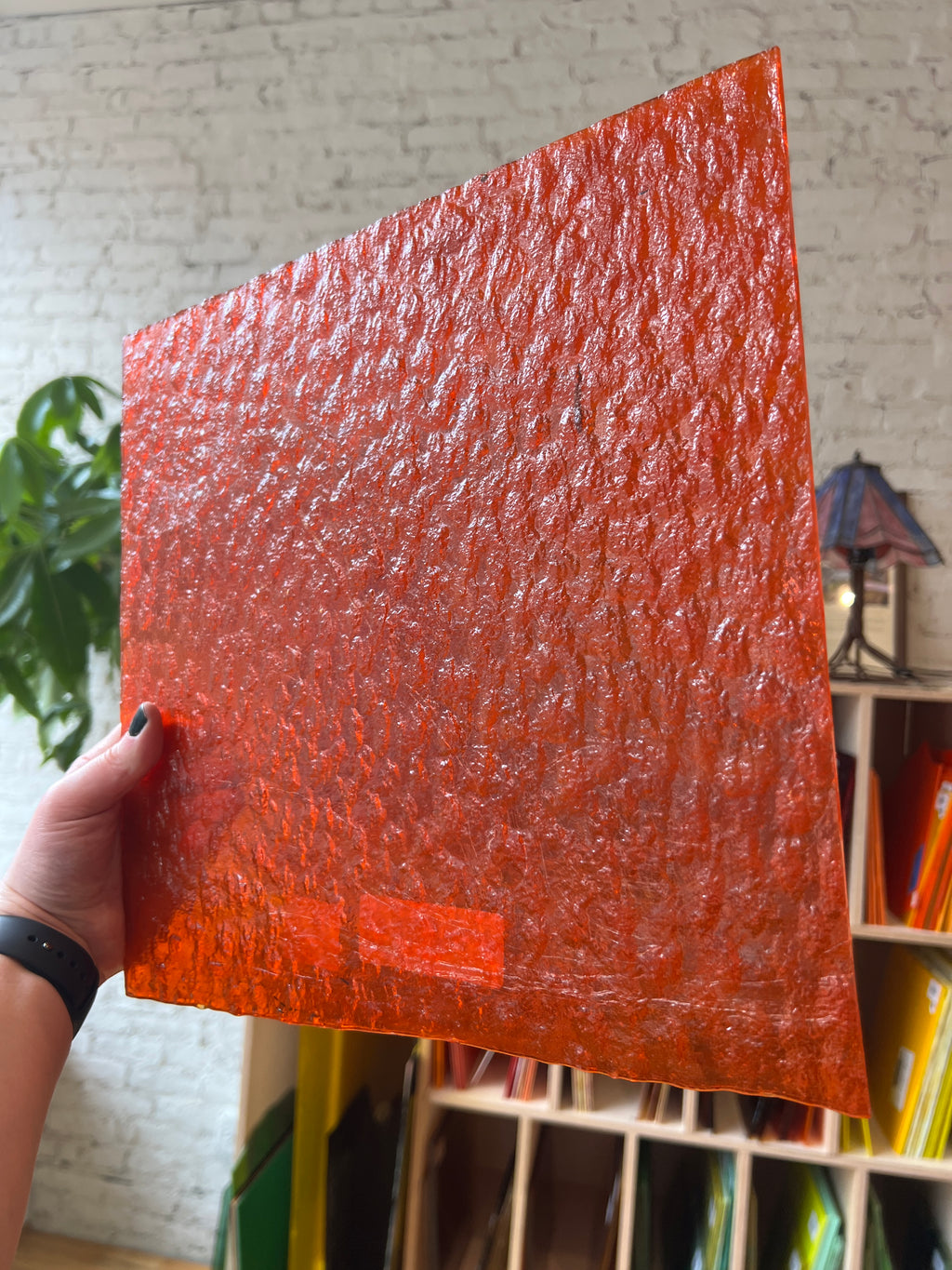 Hand holding a red textured square piece of stained glass with a blurred indoor background