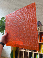 Hand holding a red textured square piece of stained glass with a blurred indoor background
