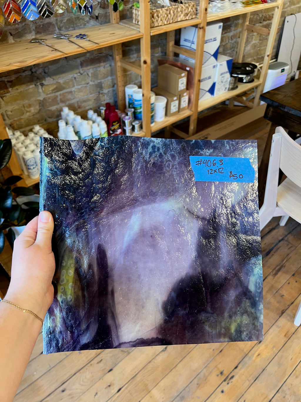 Person holding a swirled purple and white piece of stained glass in a workshop setting with shelves and tools in the background