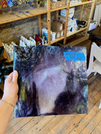 Person holding a swirled purple and white piece of stained glass in a workshop setting with shelves and tools in the background