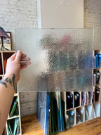 Decorative floral textured glass panel held by a hand with tattoos, surrounded by various other glass panels.