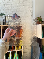 Person holding a textured rectangular sheet of stained glass with a blurred indoor background