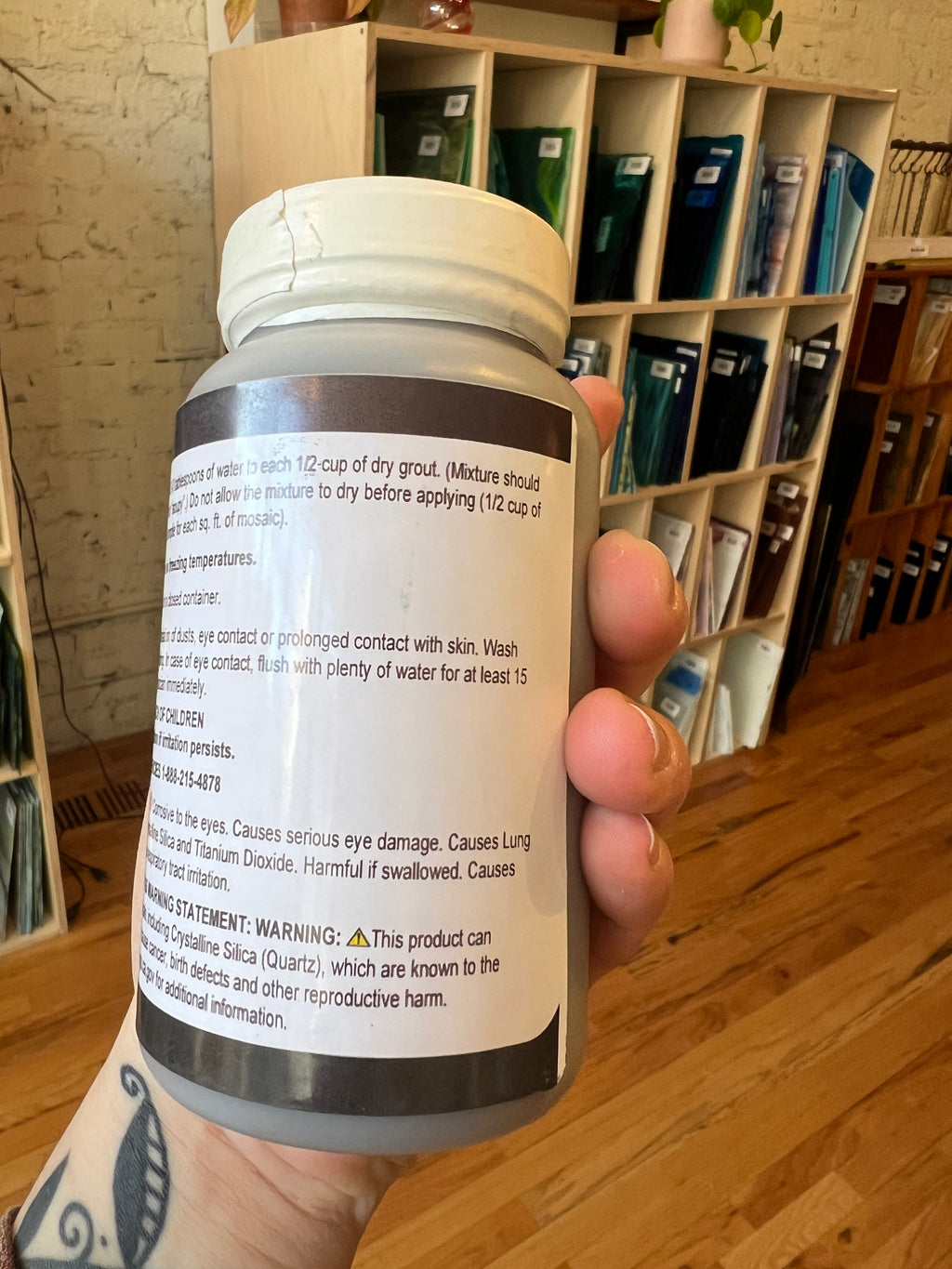 Hand holding a cylindrical container with a label, with a wooden surface and shelves in the background.