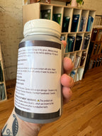 Hand holding a cylindrical container with a label, with a wooden surface and shelves in the background.