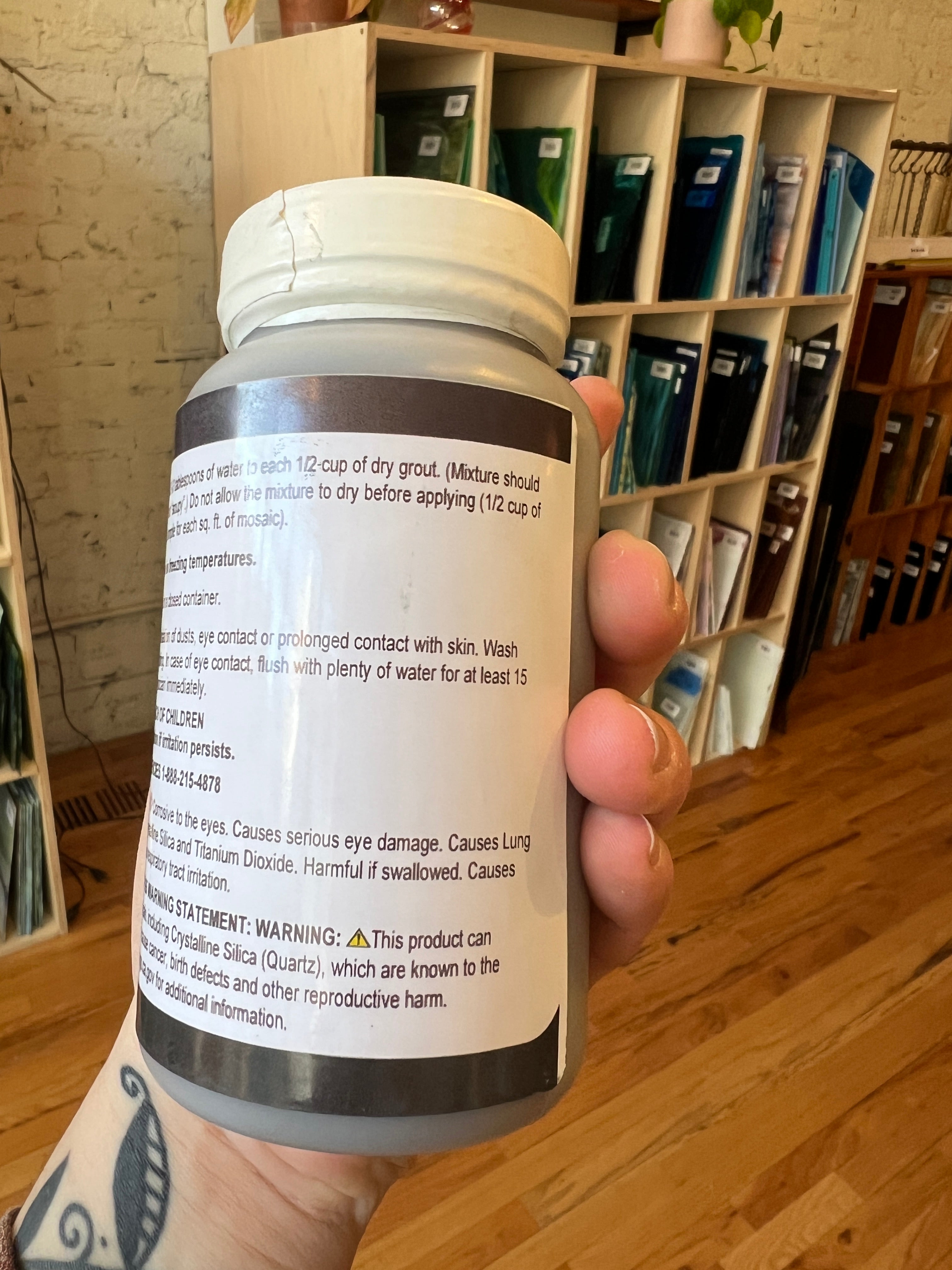 Hand holding a cylindrical container with a label, with a wooden surface and shelves in the background.