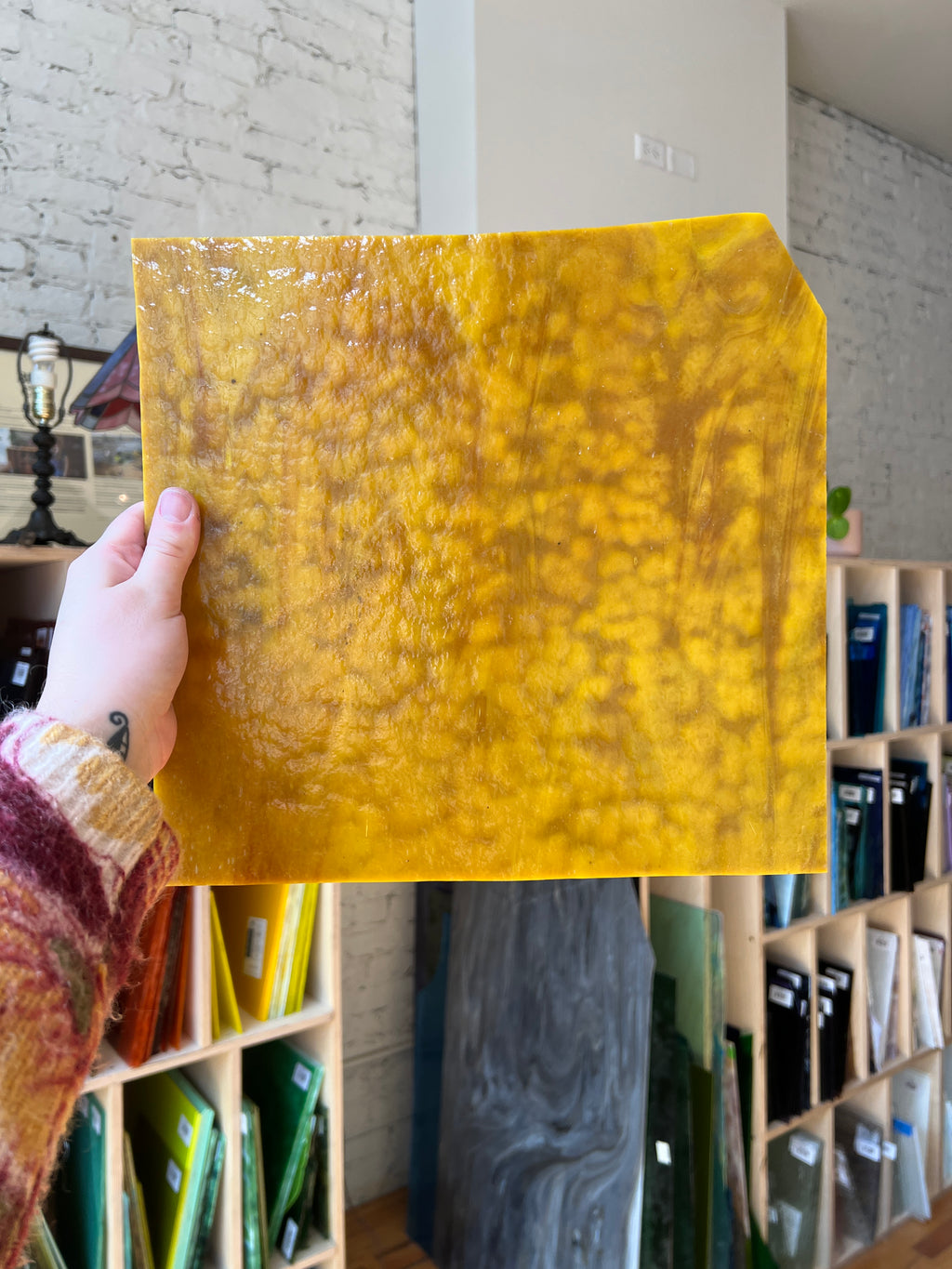 Yellow textured square of glass held by a hand with a background of various colored shelves.