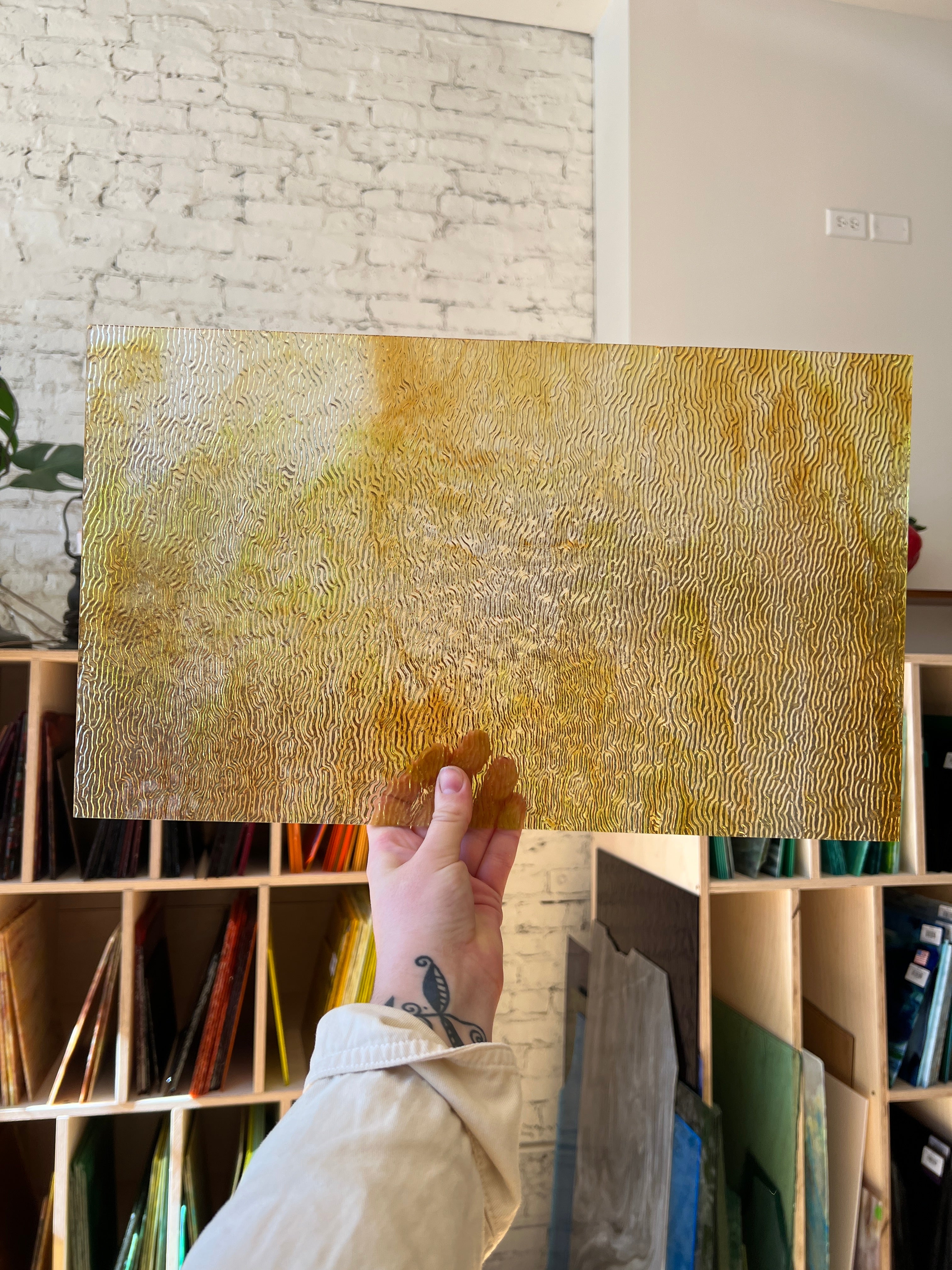 Person holding a textured gold panel of stained glass in a room with shelves and decor.