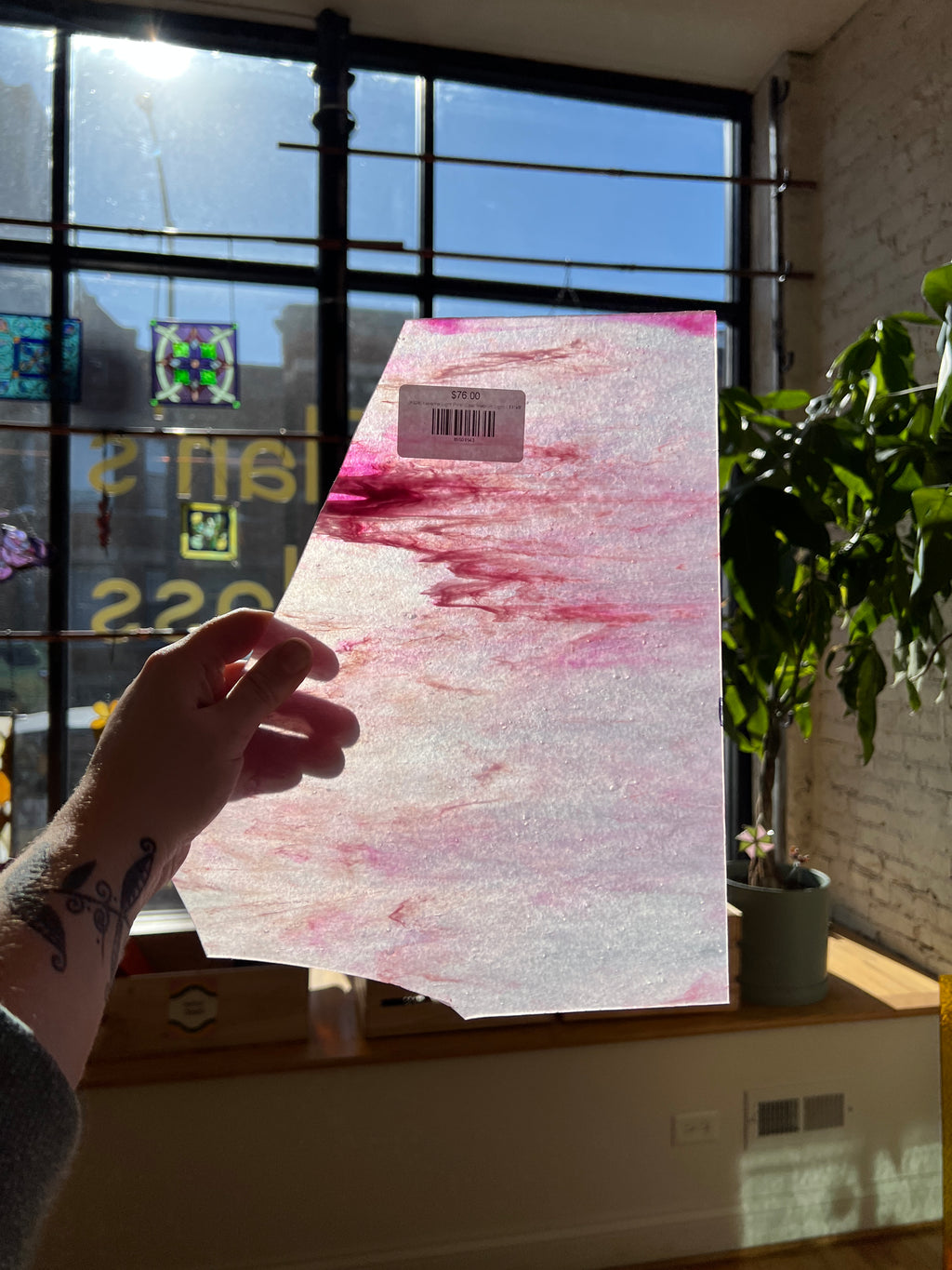 Hand holding a piece of stained glass with a pink marbled design in front of a window