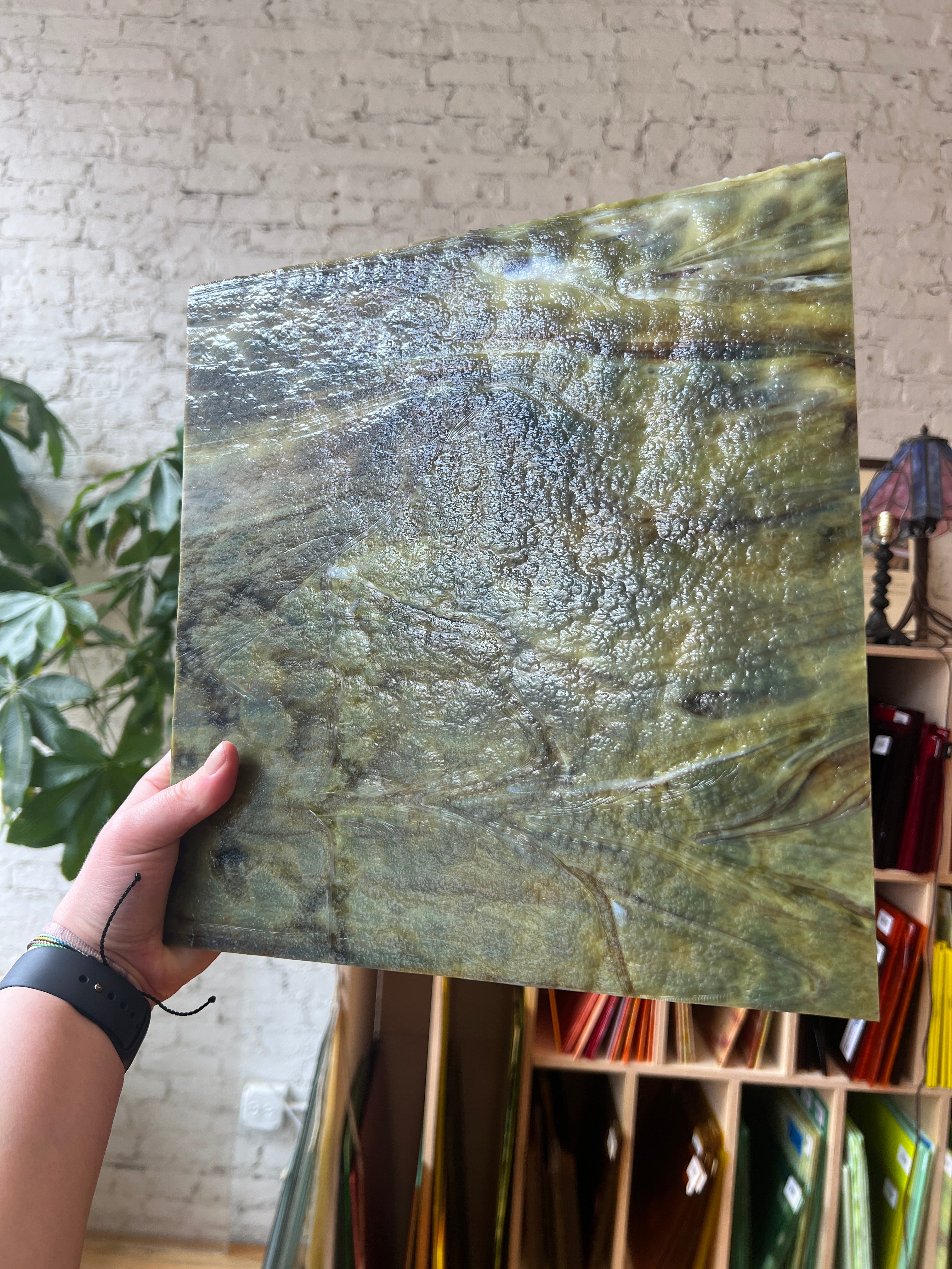 Hand holding a large green marbled sheet of stained glass against a textured wall.
