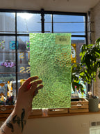 Hand holding a green textured piece of stained glass in front of a window with stained glass