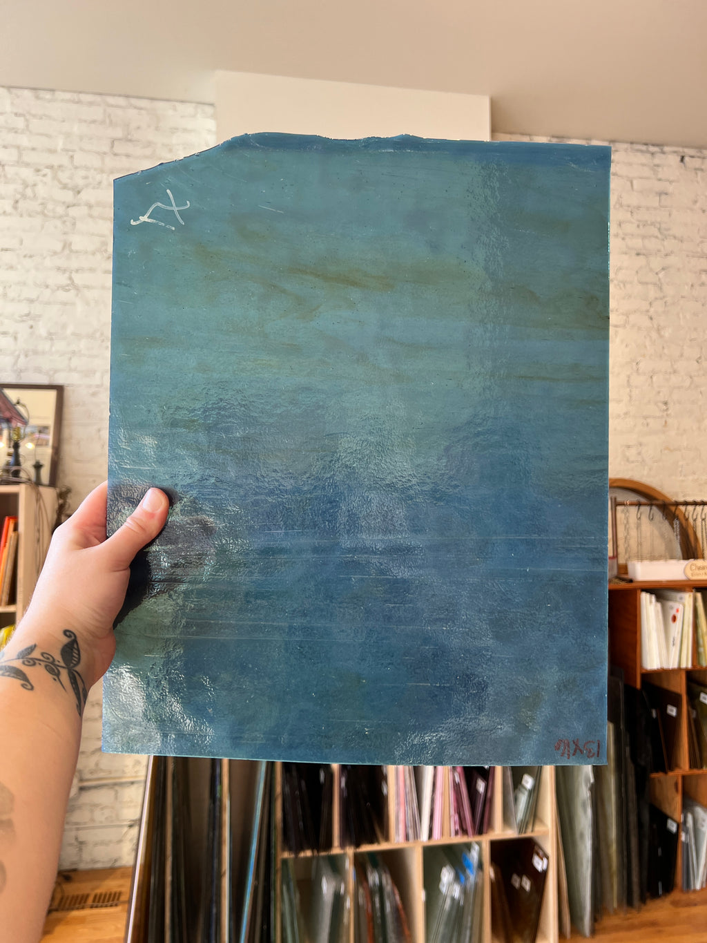 A hand with a tattoo holds up a large, textured glass sheet featuring a blue and teal gradient. The background shows a real-world art studio environment with shelves of art supplies and a white brick wall