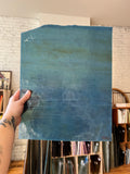 A hand with a tattoo holds up a large, textured glass sheet featuring a blue and teal gradient. The background shows a real-world art studio environment with shelves of art supplies and a white brick wall