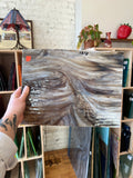 Person holding a piece of brown and white swirled textured stained glass in front of blurred shelving. 