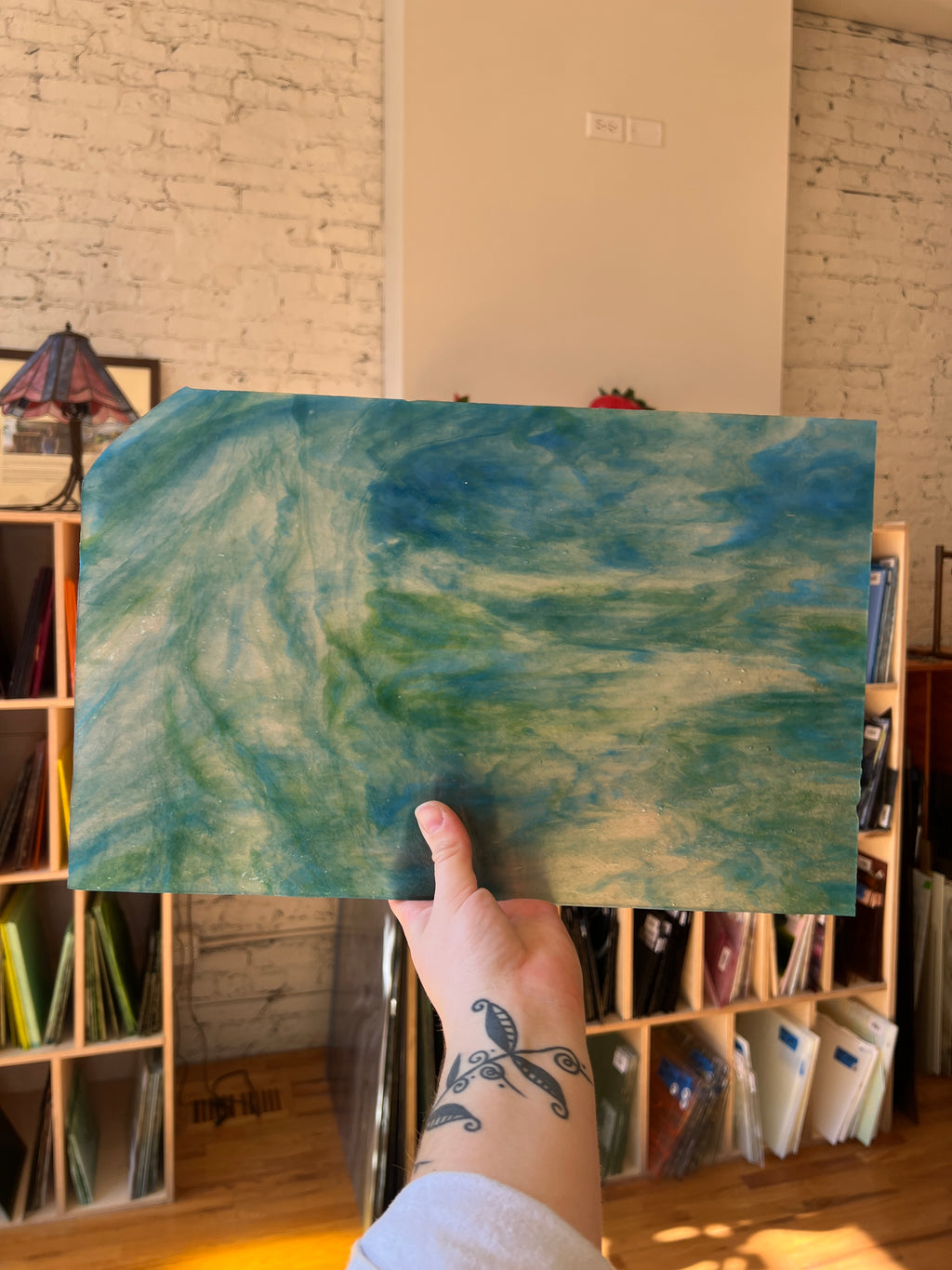 Person holding a blue and green swirled piece of stained glass in front of a blurred interior. 