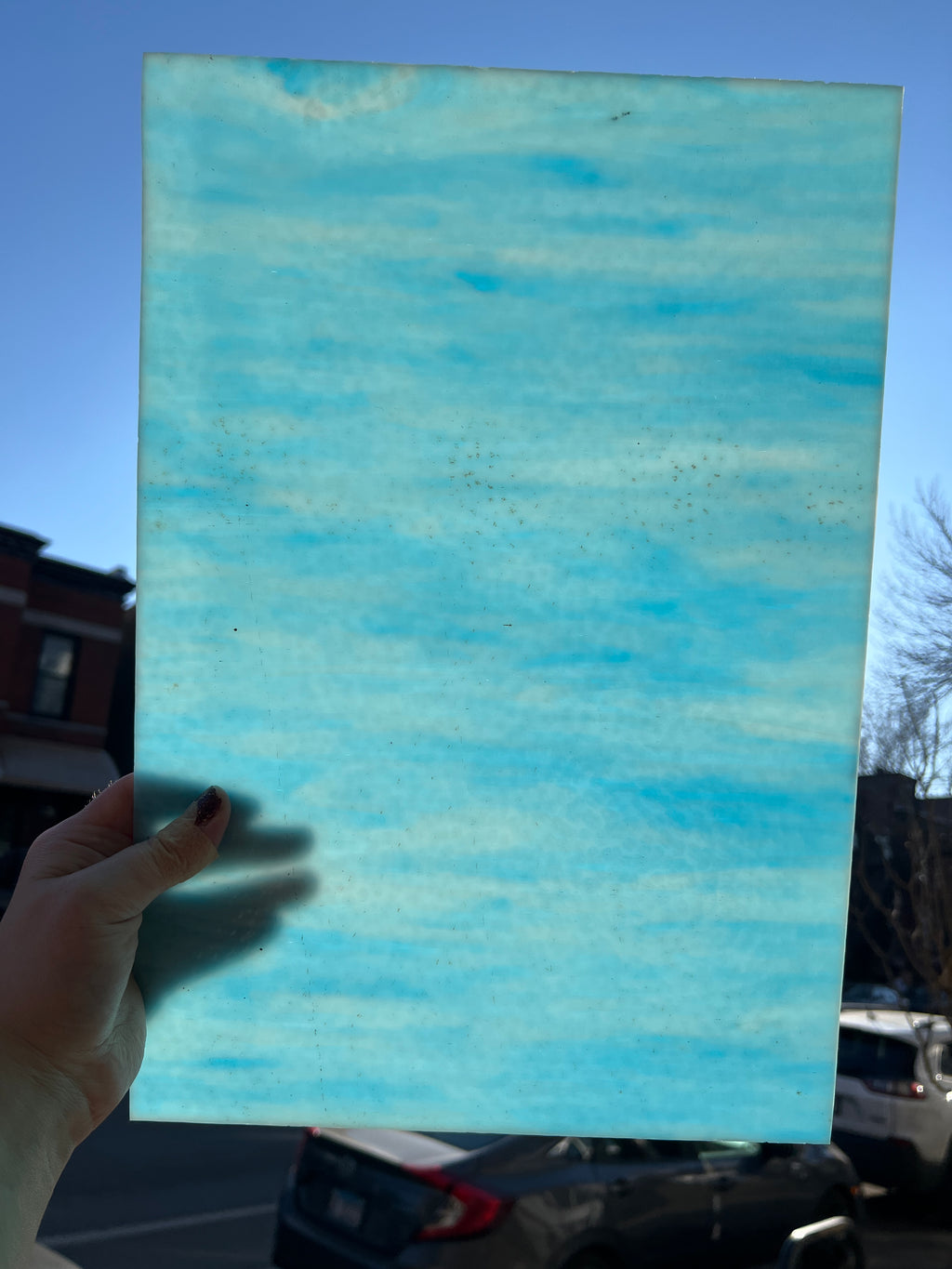 Hand holding a blue textured piece of stained glass outdoors with a clear sky.