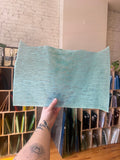 Person holding a textured light blue sheet of stained glass in front of a bookshelf.