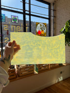 Person holding a yellowish mottled piece of stained glass in front of a blurred sunny window. 
