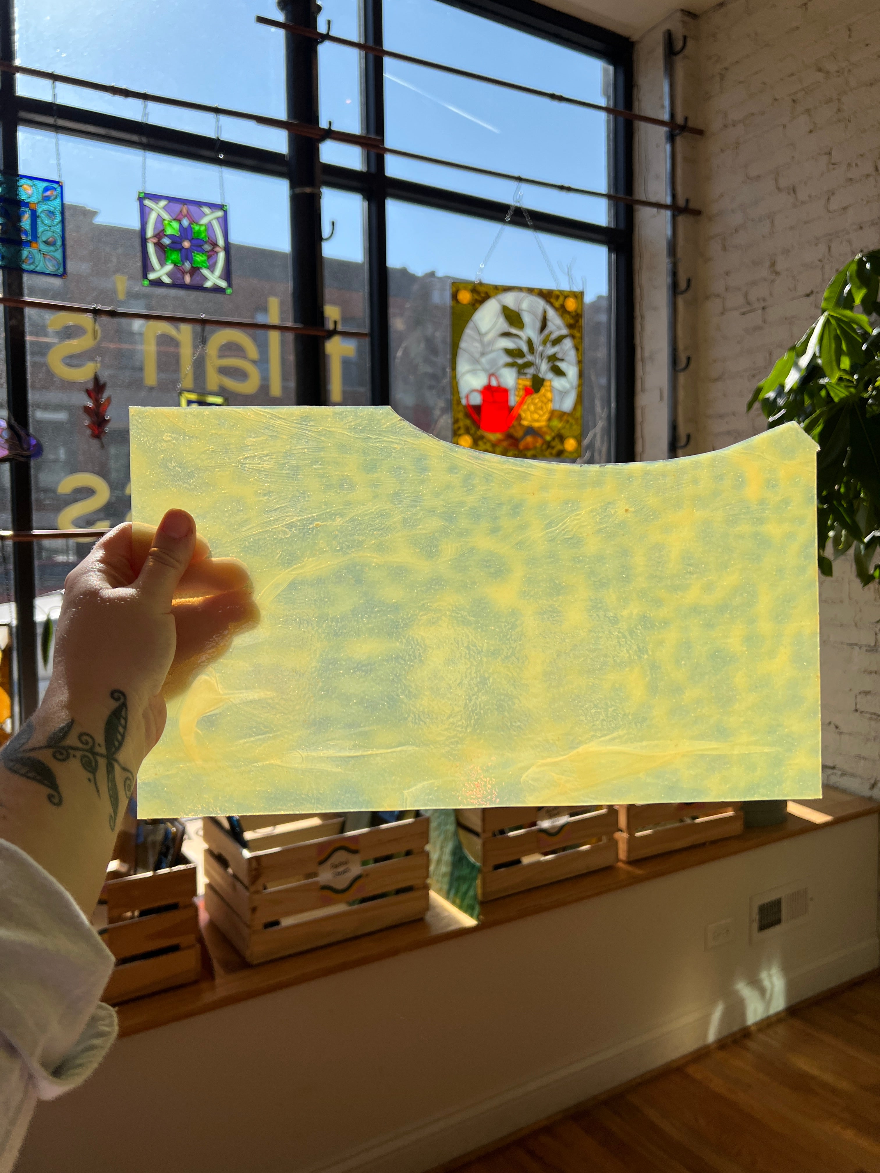 Person holding a yellowish mottled piece of stained glass in front of a blurred sunny window. 