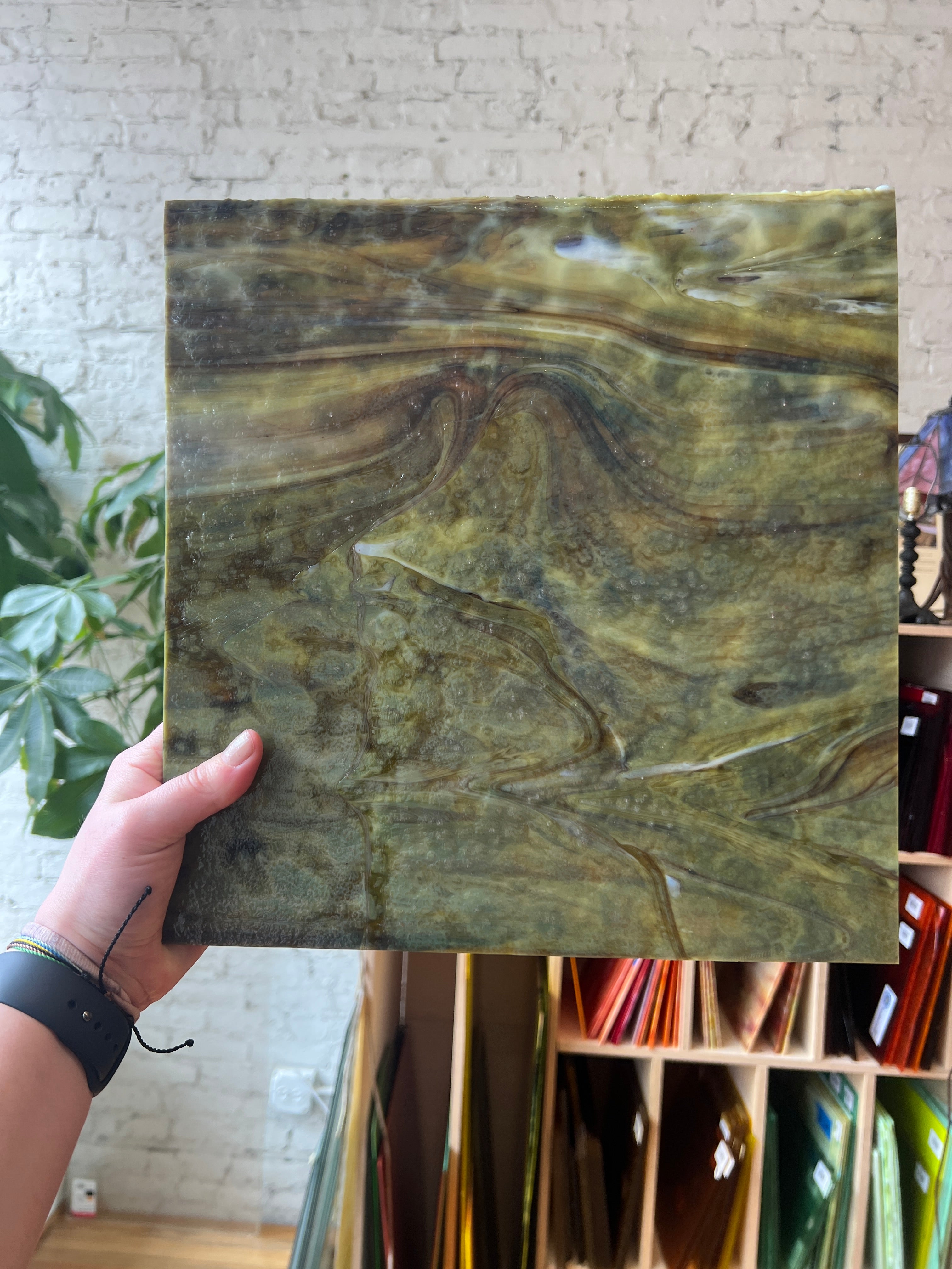 Hand holding a green marbled sheet of stained glass against a textured wall.