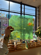 Person holding a green textured piece of stained glass in front of a blurred sunny window. 