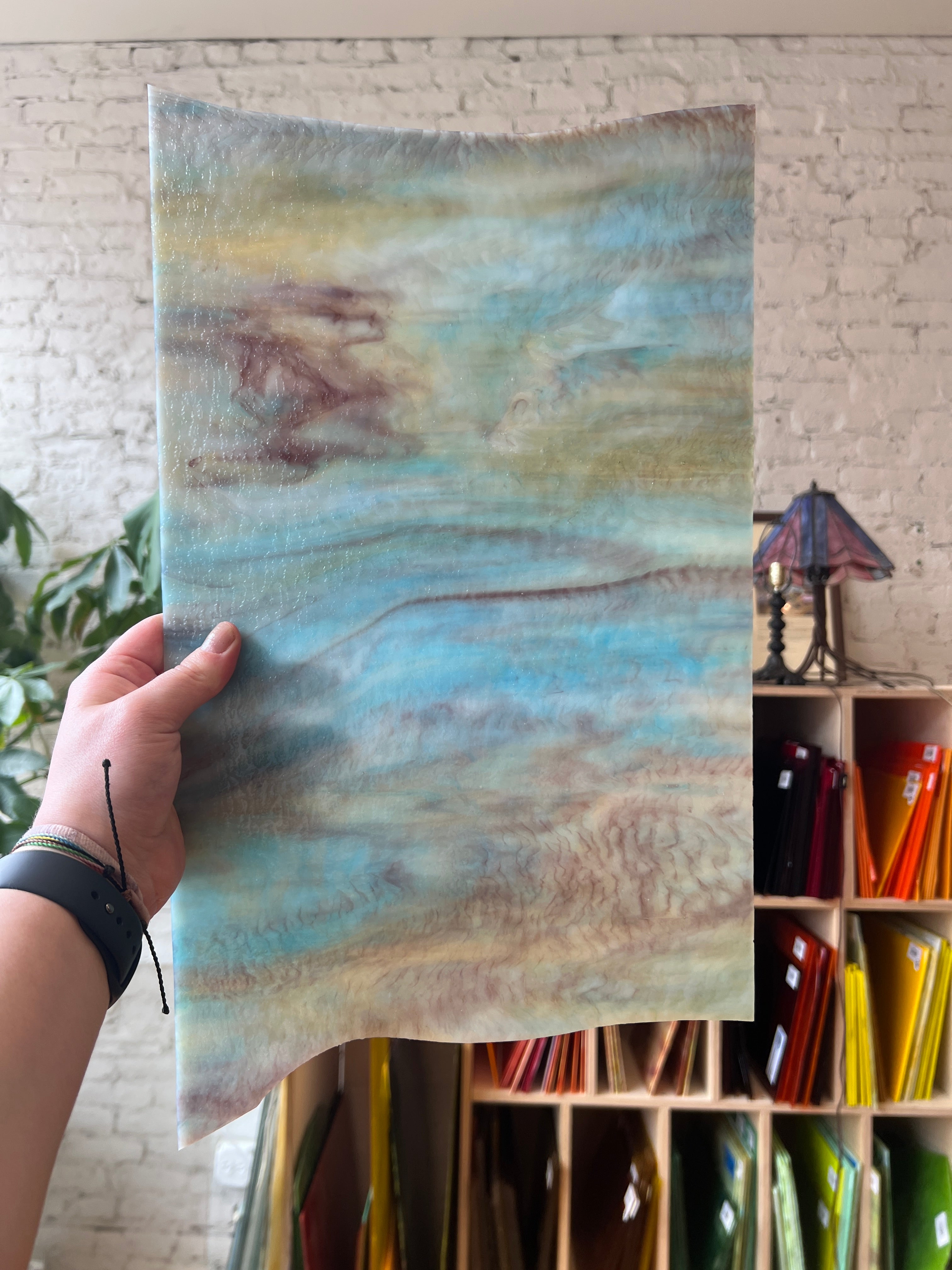 Hand holding a piece of glass with a watercolor-like pattern in front of a colorful shelf.