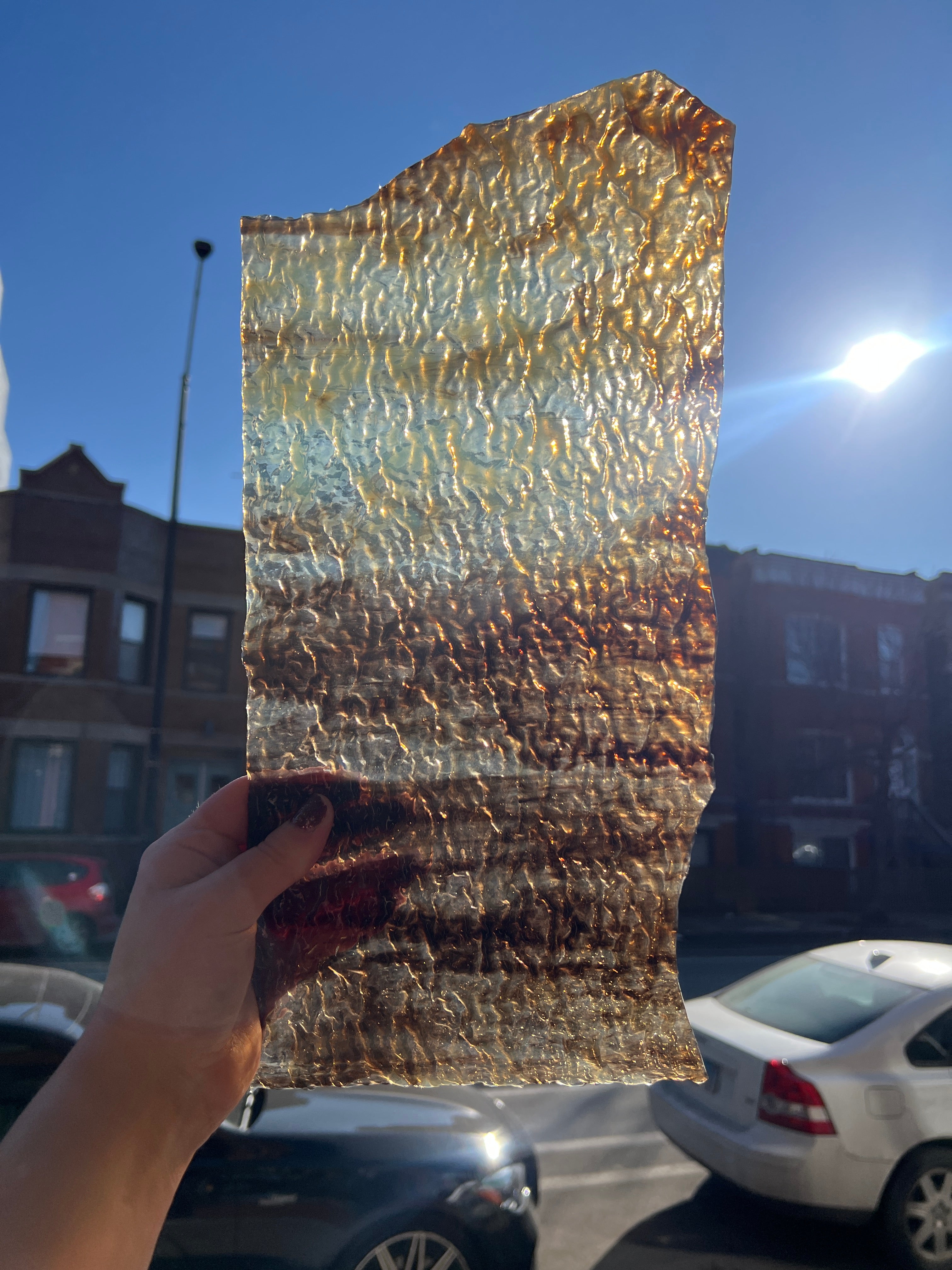 Person holding a piece of textured streaky brown and white stained glass against a blue sky with buildings and a car in the background.