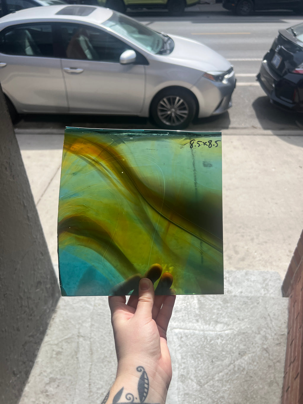 Hand holding a square piece of stained glass with blue and green colors on a street background.