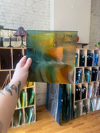 Hand holding a piece of stained glass with a blurred background of shelves with vases.